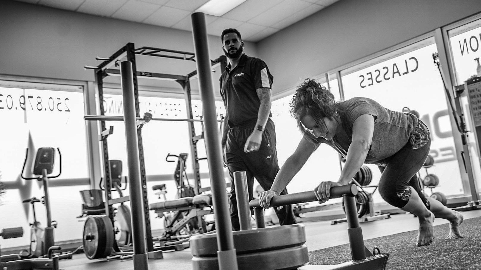 A client drives a weighted sled while Cassidy coaches from beside the rack